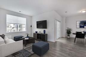 A living room with a white couch, a black chair, a black ottoman, a flat screen TV, and a window with a view of a building outside.