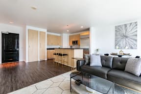 A black couch sits in a living room with a wooden floor and a bar area in the background.