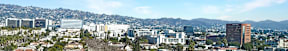 A cityscape with buildings and trees in the foreground and mountains in the background.