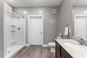 Shawnee KS Pet-Friendly Luxury Apartments - Bonavia Luxury Apartments - Bathroom with Vinyl Plank Flooring, Vanity Mirror, Sink, Dark Brown Cabinetry, and Open Shower.