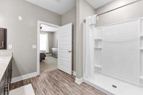 A white bathroom with a walk-in shower and a bedroom visible through the door.