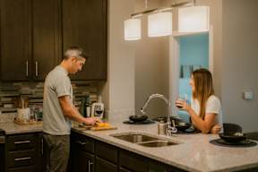 A man and a woman are in a kitchen, the man is preparing food and the woman is drinking from a cup.