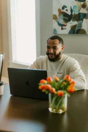 A man is sitting at a table with a laptop and a vase of flowers.