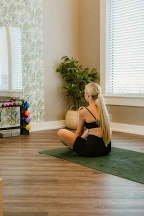 A woman with blonde hair is sitting on a green mat in a room with a plant and a window.