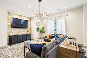 A living room with a grey couch, a wooden chessboard, and a television on the wall.