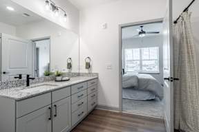 A bathroom with a mirror, sink, and a bedroom visible through the door.