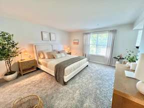 A bedroom with a large bed, a nightstand with a lamp, a plant, and a window with curtains.