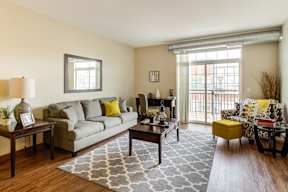 Osseo, MN Studios for Rent | 5 Central | Living Room with Wood-Style Flooring, Area Rug, Sofa, Coffee Table, End Table with Lamp, Armchair, Stool, Desk with Chair, and Sliding Glass Door with Balcony Access.