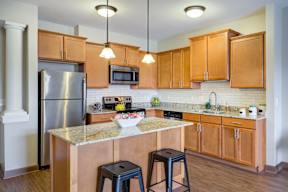 Studio Apartments in Osseo, MN | 5 Central | Kitchen with 9-foot Ceiling, Stainless Steel Appliances, Tile Backsplash, Wood-Tone Cabinetry, Kitchen Island with Seating, Pendant Lights, and Wood-Style Flooring.