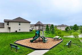 Townhomes in Peculiar, MO - Bradley’s Crossing - An Outdoor Playground Featuring a Play Structure on a Wood Fiber Surface Surrounded by Grass, Trees, Benches, and Townhomes.