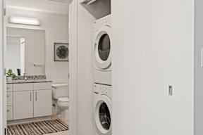 A white washing machine is in a laundry room with a toilet and sink in the background.