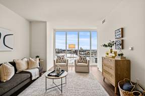 A living room with a grey couch, a coffee table, and a large window with a view of the city.