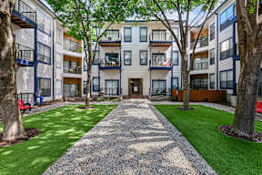 A gravel pathway leads to a building with balconies and trees on either side.