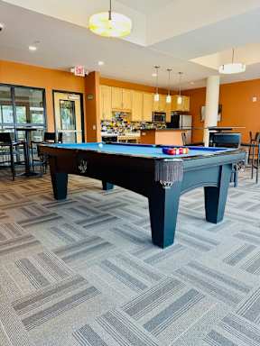 A spacious recreation room here at Gallery Flats Apartment Homes with a blue felt billiard table centered, surrounded by modern furniture, orange walls, and a kitchen area in the background.