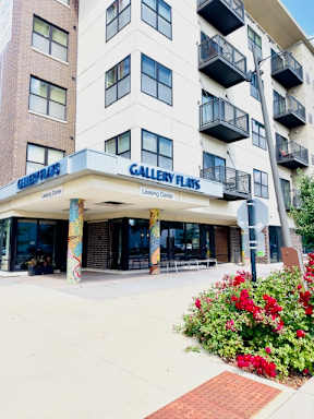 Gallery Flats apartment building with a beige facade, black balconies, and colorful pillars. Bright flowers in the foreground create a welcoming atmosphere.