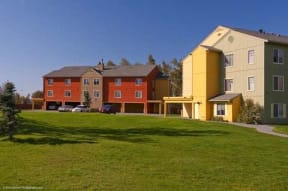 Colorful townhouses here at Brighton Park with red, yellow, and green facades stand against a clear blue sky, surrounded by lush green grass and a few trees.