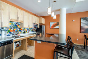 Modern kitchen here at Gallery Flats Apartment Homes with light wood cabinets, vibrant mosaic backsplash, and stainless steel appliances. Orange walls, bar counter with stools, and warm lighting.