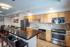 Modern kitchen here at Gallery Flats Apartment Homes with light wood cabinets, stainless steel appliances, and mosaic tile backsplash. A granite island with bar stools and dining area is visible.
