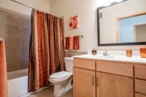 A cozy bathroom here at Gallery Flats Apartment Homes with warm tones featuring a large mirror, wooden vanity, and a striped brown and orange shower curtain. Decor includes orange accents.