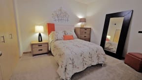 Cozy bedroom here at Gallery Flats Apartment Homes with a floral-patterned bed, wooden nightstand, lamps, and a large mirror. Warm lighting creates a comfortable, inviting atmosphere.