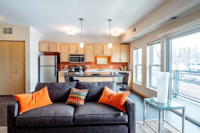 Modern living room and kitchen here at Gallery Flats Apartment Homes in an open layout. A gray sofa with orange cushions faces a kitchen with wooden cabinets. Large windows brighten the space.