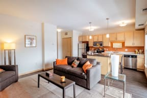 Modern open-concept living space here at Gallery Flats Apartment Homes with beige walls. Features a gray sofa with orange and patterned pillows, a sleek coffee table, and a cozy floor lamp. Adjacent kitchen with light wood cabinets, stainless steel appliances, and warm ambient lighting. Large windows create a bright, inviting atmosphere.
