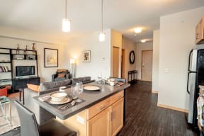 Modern, cozy apartment living room here at Gallery Flats Apartment Homes with open kitchen. Features a gray sofa, dining setup on a kitchen island, and neatly arranged shelves, conveying warmth.