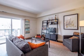 Modern living room here at Gallery Flats Apartment Homes with gray sofas, orange cushions, and a black coffee table. A TV sits on a bookshelf. Large window, bright and cozy ambiance.