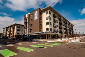 Street view of our modern Gallery Flats Apartment Homes building with balconies, marked "Now Renting," under a blue sky. Green crosswalk lines dominate the foreground.