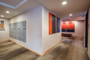 A modern hallway here at Gallery Flats Apartment Homes building with gray mailboxes on the left wall, abstract red and blue paintings on the right, and soft lighting creating a calm ambiance.
