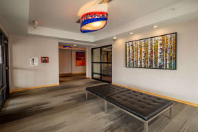 Modern hallway here at Gallery Flats Apartment Homes with a black leather bench, abstract tree artwork on the wall, and colorful ceiling light, creating a contemporary, artistic atmosphere.