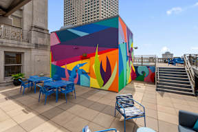 A colorful mural on a building with blue chairs and tables in front.