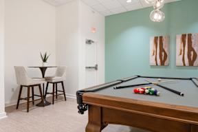 A pool table with a few balls on it and a small table with chairs in the corner.