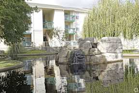 A large rock formation sits in the middle of a pond in front of a white apartment building.