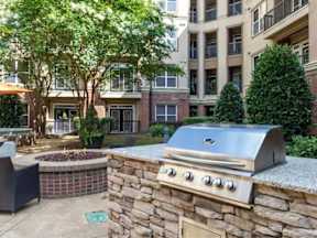 A stone BBQ grill is in the middle of a patio with a fire pit and chairs.