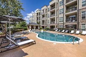 A large swimming pool surrounded by lounge chairs and umbrellas in front of apartment buildings.