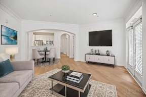 A living room with a grey couch, a black coffee table, a television, and a dining area in the background.