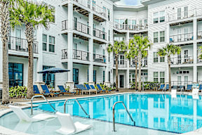 A swimming pool with lounge chairs and palm trees in front of a white apartment building.
