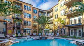 A swimming pool is surrounded by lounge chairs and palm trees in front of apartment buildings.