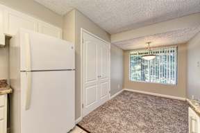 A white refrigerator stands in a kitchen with a carpeted floor and a window with blinds.