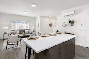 A modern kitchen with a white countertop and dark wood cabinets open to the living room with a couch and two sitting chairs.