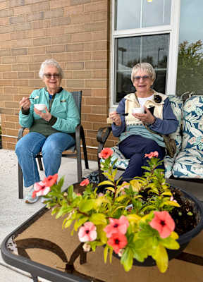 Two older women sitting outside on chairs with a planter in front of them.