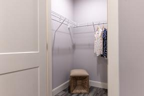 A small closet with clothes hanging on a wire rack and a stool in front.