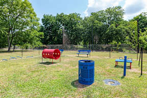 A park with a red and blue playground structure, a blue trash can, and a fence.