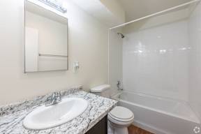 A bathroom with a marble countertop, a white toilet, a sink, a mirror, and a tub with a shower.