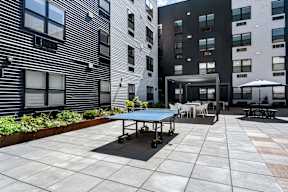 A blue table tennis table is surrounded by chairs and umbrellas in a courtyard.