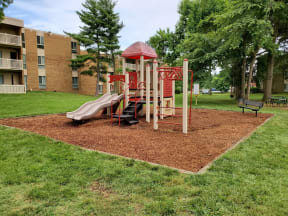 a playground with slides and monkey bars in a grassy area with a brick building in the at The Glendale Residence, Maryland, 20706
