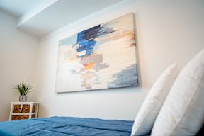 A large abstract painting hangs on a white wall above a bed with blue sheets.