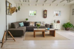 Living room with white brick walls and light hardwood floors. There is a sectional couch in left corner and a modular coffee table in the center of the room. There are two windows on the back wall and one window out of frame on the right. The ceiling is sloped slightly with support beams along its length.