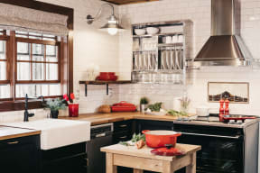 Kitchen with white tile walls, stainless steel fixtures and appliances, light wood counters, and black cabinets. The oven is on the right with a large fume hood. On the left is a window above the sink.